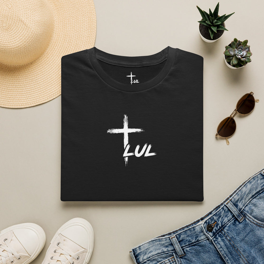 A folded black T-shirt with a white cross and the word 'LUL' printed on the front, styled in a flat lay with a straw hat, small potted succulents, sunglasses, white sneakers, and blue jeans on a light beige background.
