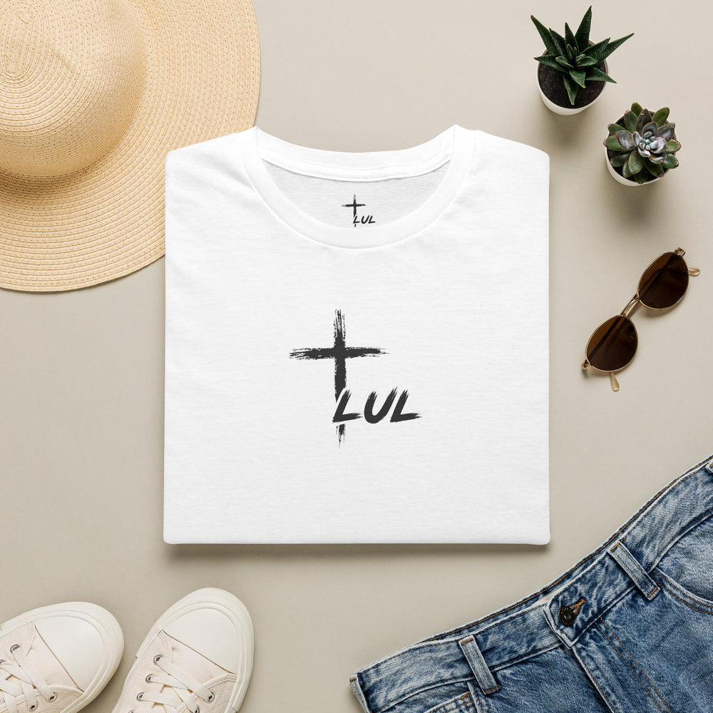 White t-shirt with a black geometric design, placed on a light surface with a hat, sunglasses, and jeans.
