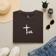 A folded dark brown t-shirt with a white graphic reading 'LUL' under a cross, styled in a flat lay with a straw hat, white sneakers, sunglasses, a pair of blue jeans, and small potted succulents on a light beige background.
